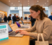 Voici le texte alternatif (SEO/accessibilité) ainsi que la description détaillée de l'image finalisée : Texte alternatif (ALT) Femme utilisant un MacBook dans un Apple Store près d'un présentoir de sondage sur les picotements électriques.